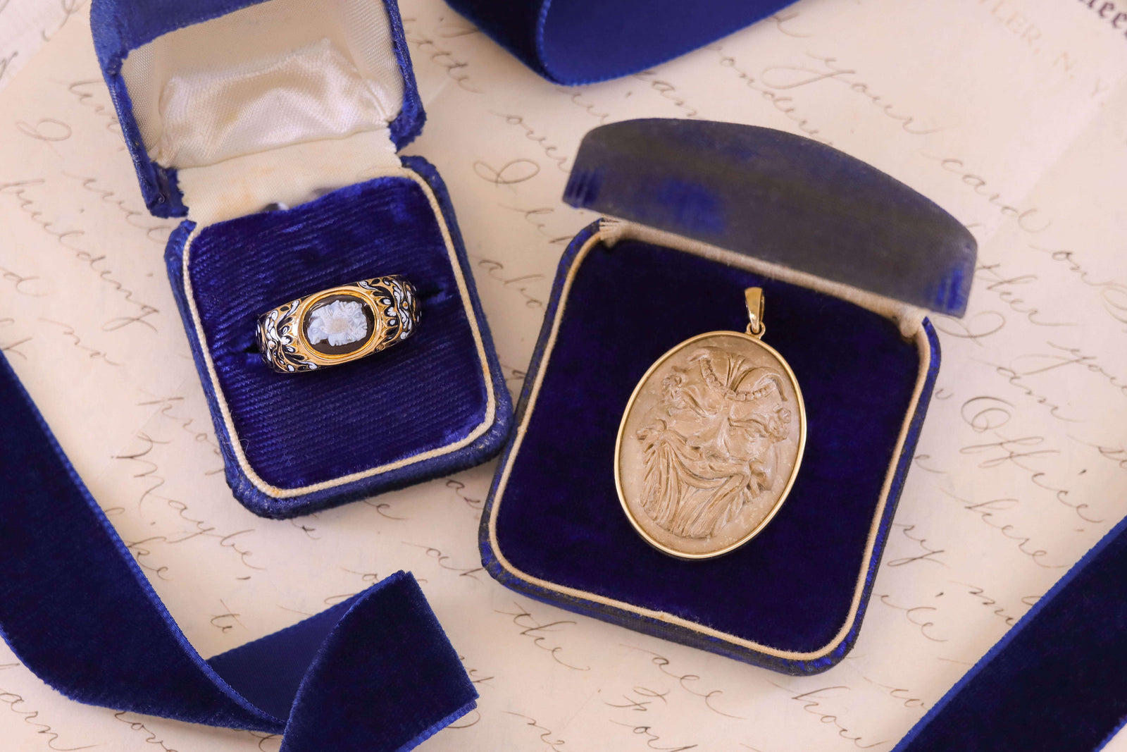 Antique cameo ring and pendant displayed in blue velvet boxes on handwritten paper. The ring features a four faced profile carved in stone, framed by black and white enamel scrollwork; the pendant shows a three faced man carved in lava, set in gold.