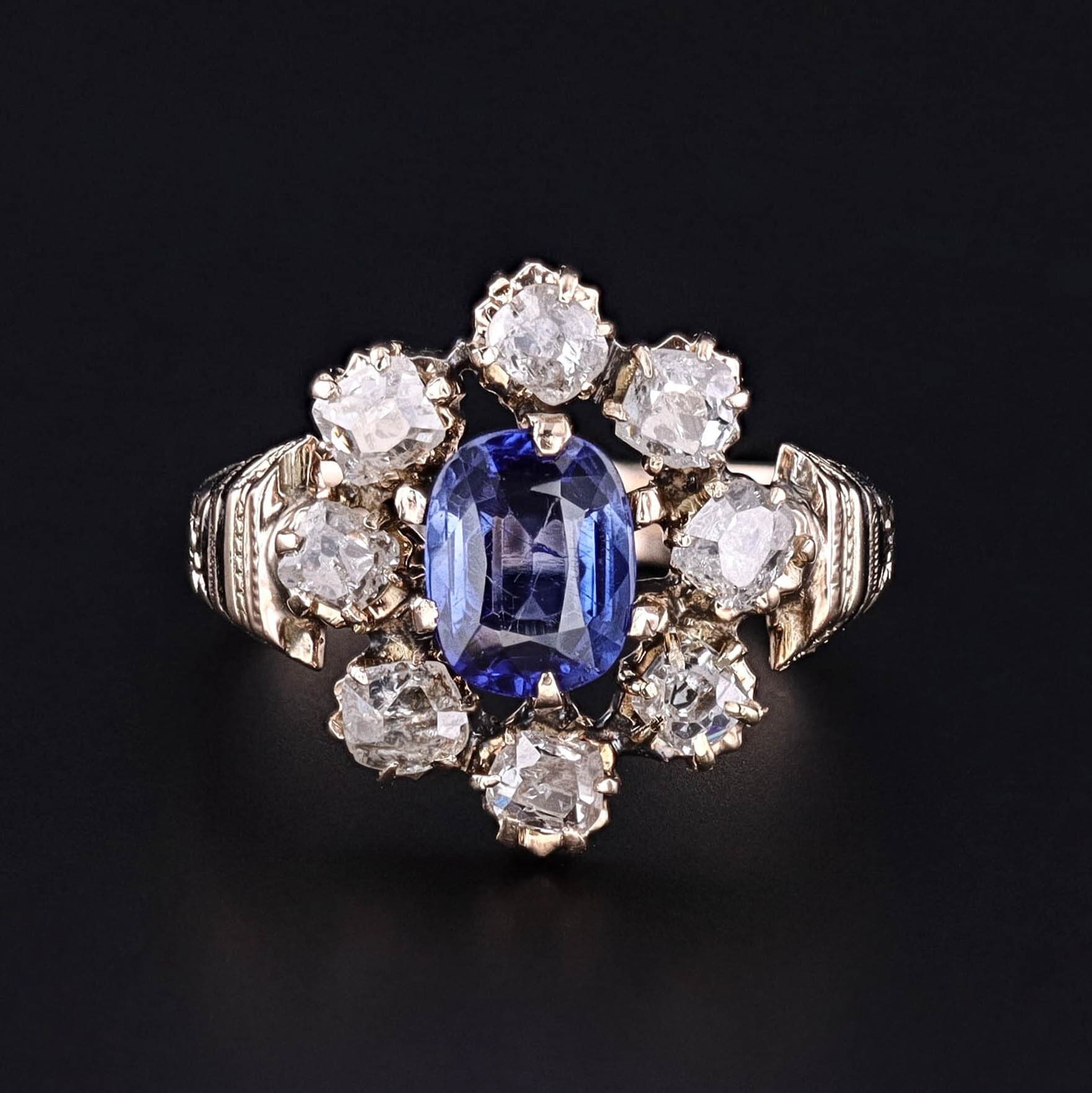 A late Victorian era ring (circa 1880-1900), showcasing a striking synthetic blue sapphire encircled by a halo of old mine cut diamonds. The ring is 12ct gold with black enamel accents.