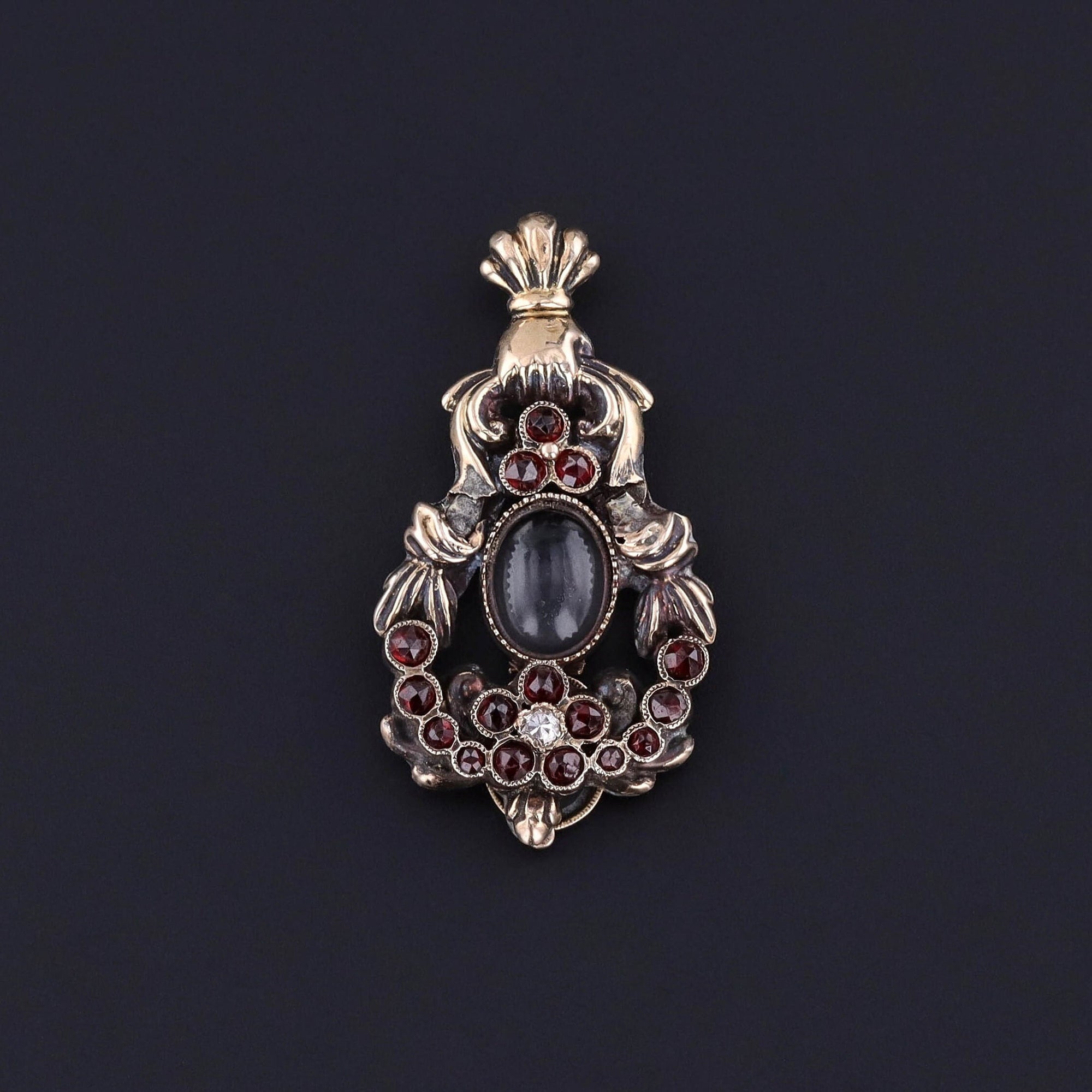 Transport yourself back in time to the Georgian era with this antique brooch conversion. The pendant features a hand holding a garland of rose cut garnet flowers with an oval glass locket at its center and a diamond accent.