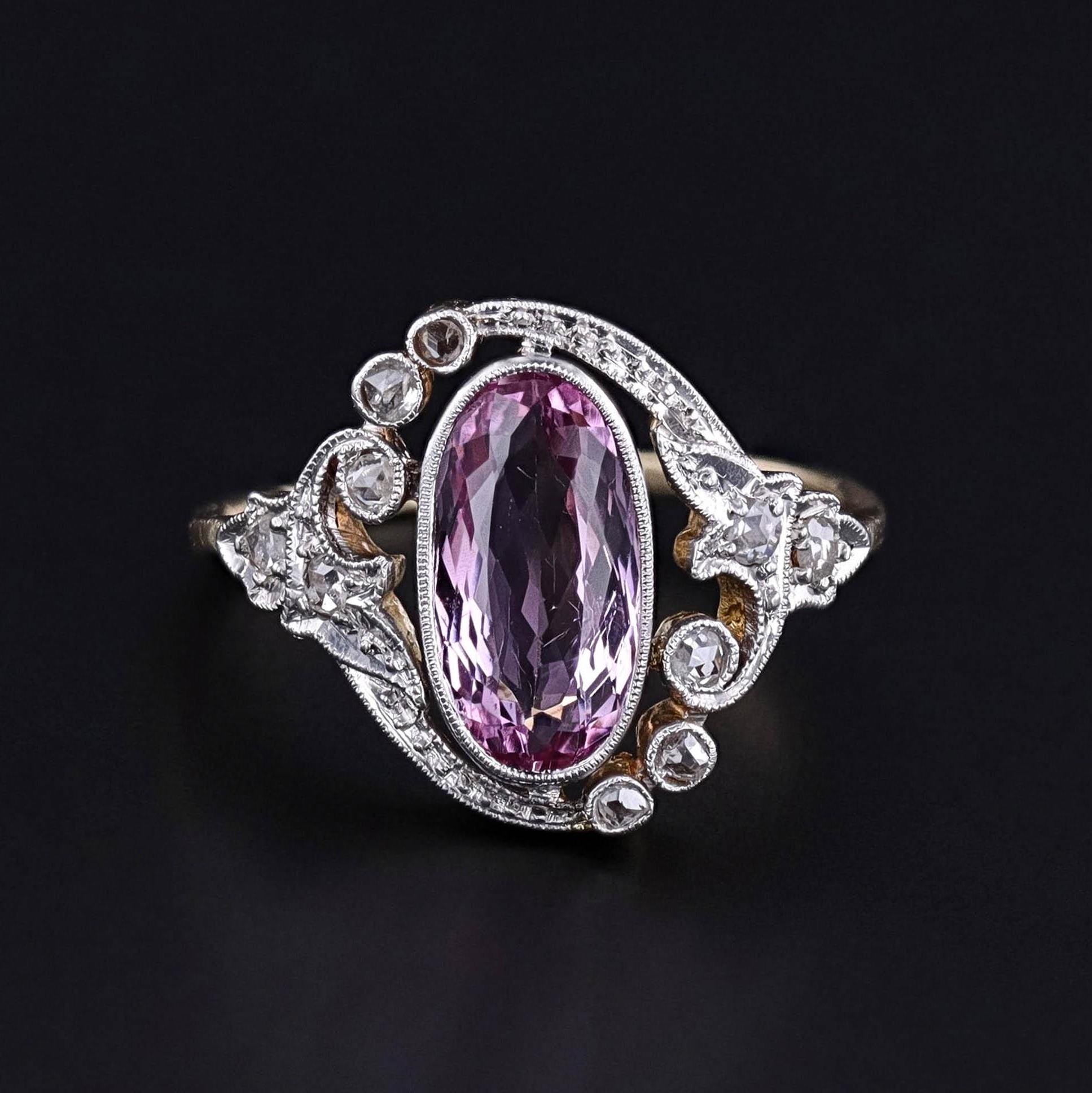 The image shows a Belle Epoque ring featuring an oval-cut pink topaz at its center, surrounded by intricate detailing of diamonds and scrolling metalwork. The antique ring&#39;s design includes round diamonds on both sides, enhancing its elegance.