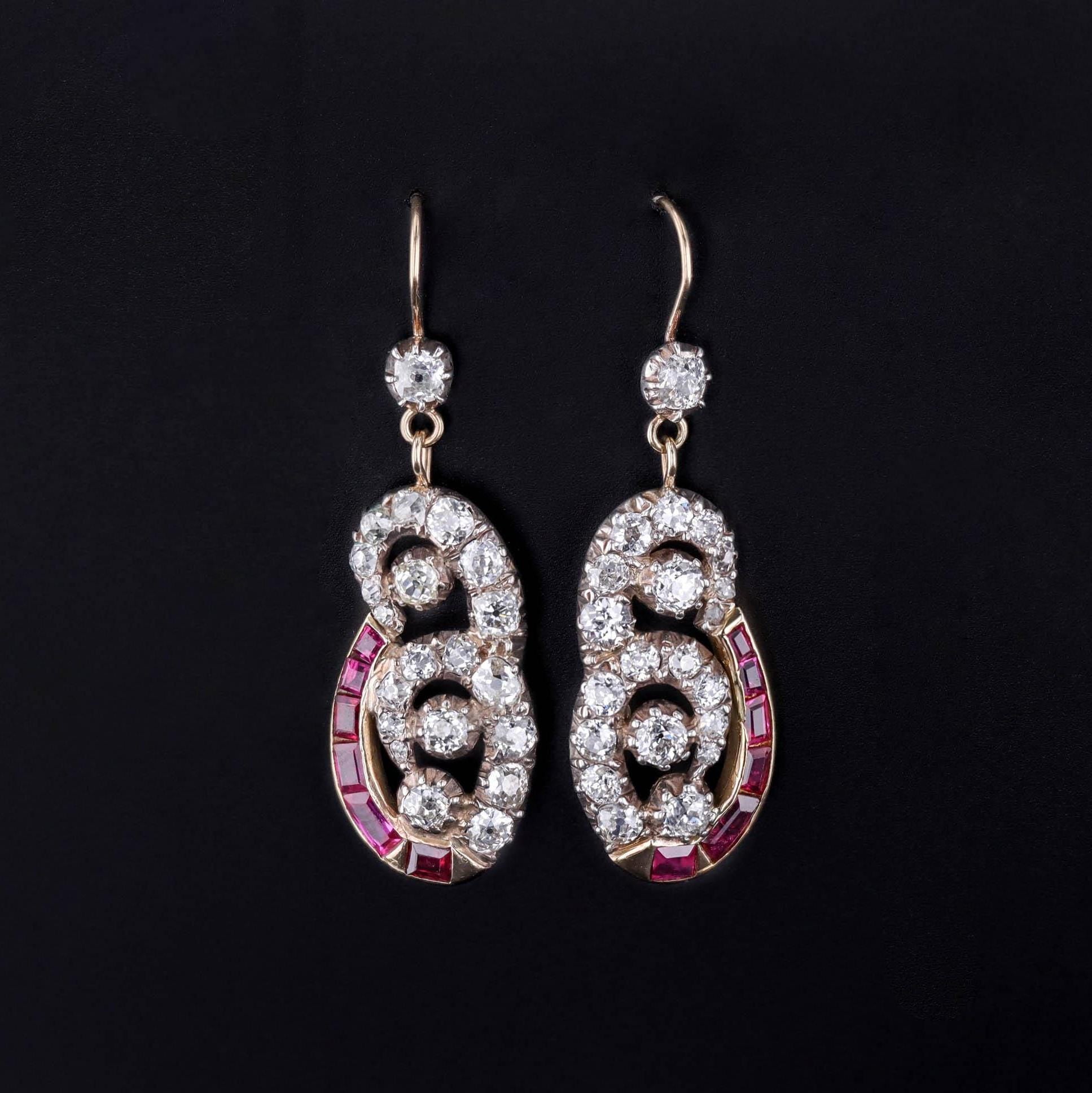 The image shows a pair of vintage-style drop earrings with swirling designs adorned with round-cut diamonds and a row of square-cut rubies. A single diamond connects the hooks to the intricate lower section.